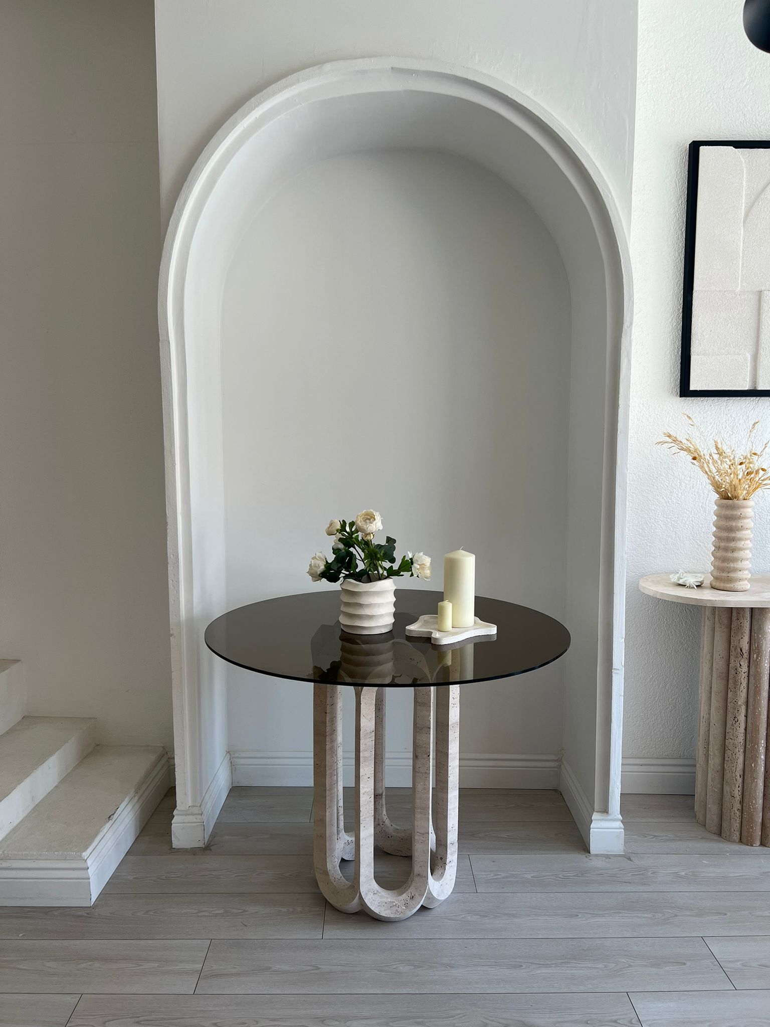 Modern interior featuring Luzione Eos glass top travertine dining table and cylindrical travertine side table. Minimalist decor with decorative arch