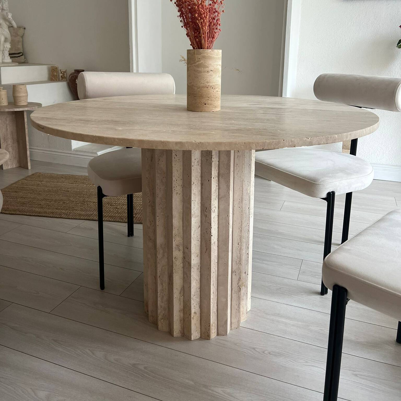 Interior view of a dining area featuring a round travertine dining table with a fluted base, surrounded by modern chairs with light upholstery