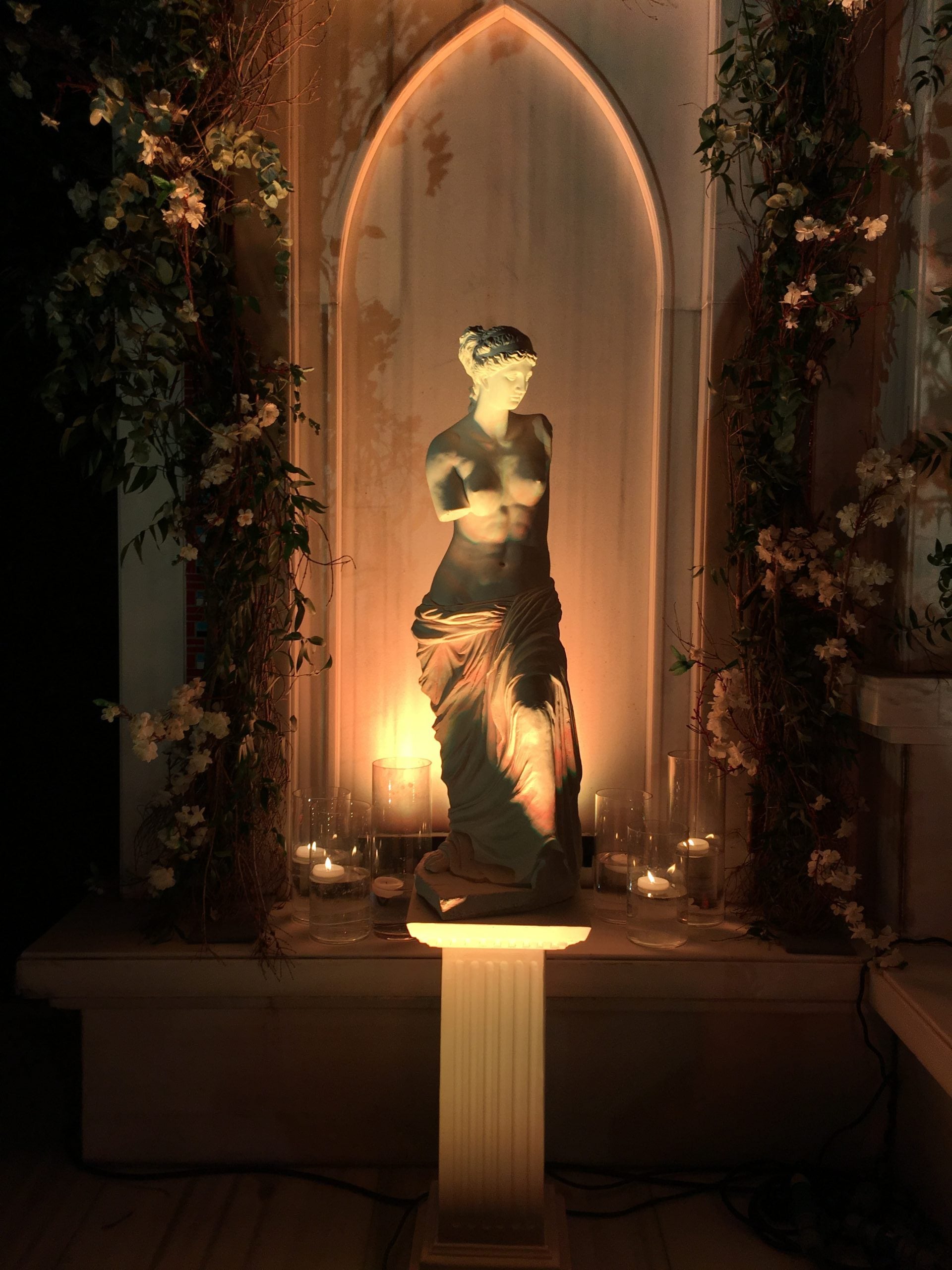 Nighttime view of white Venus de Milo statue in arched niche, lit from below.