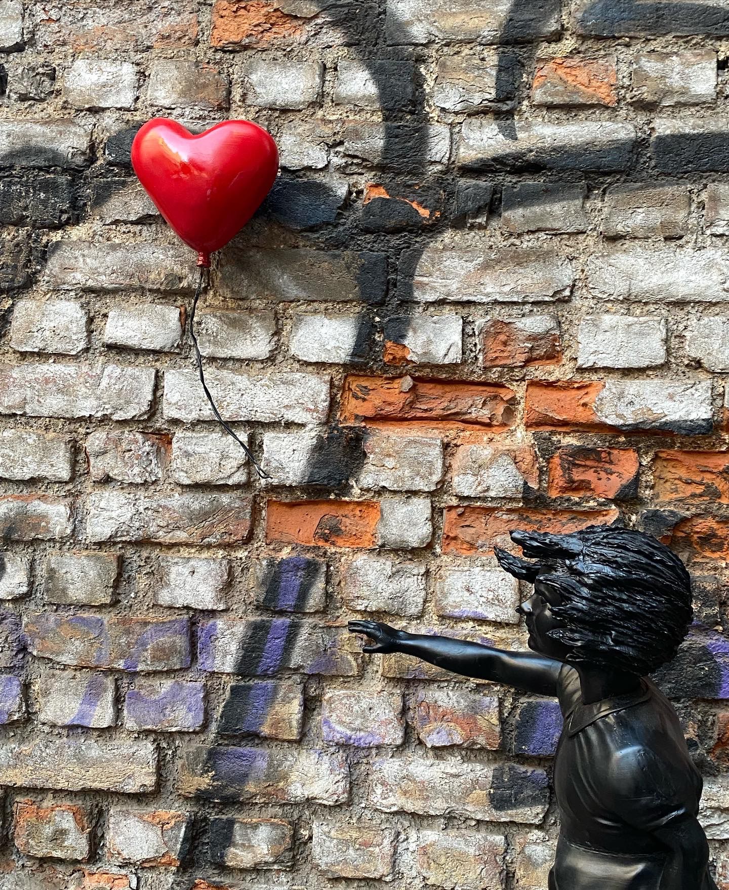 Black child statue reaching for red heart balloon against brick wall