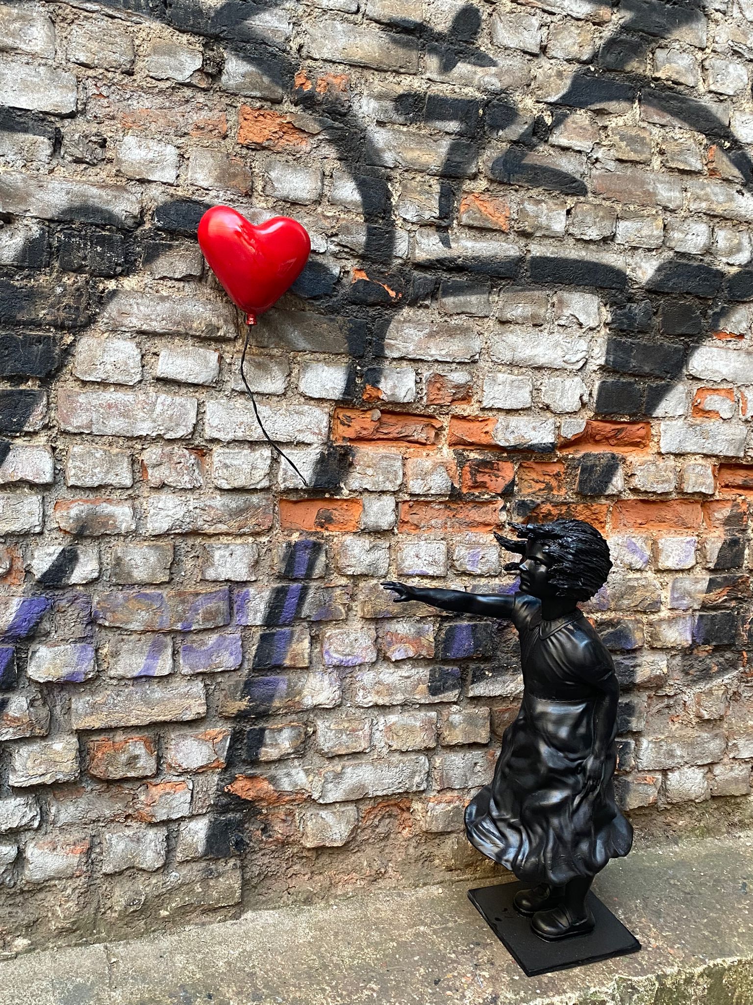 View of black girl statue pointing at red heart balloon on brick wall.