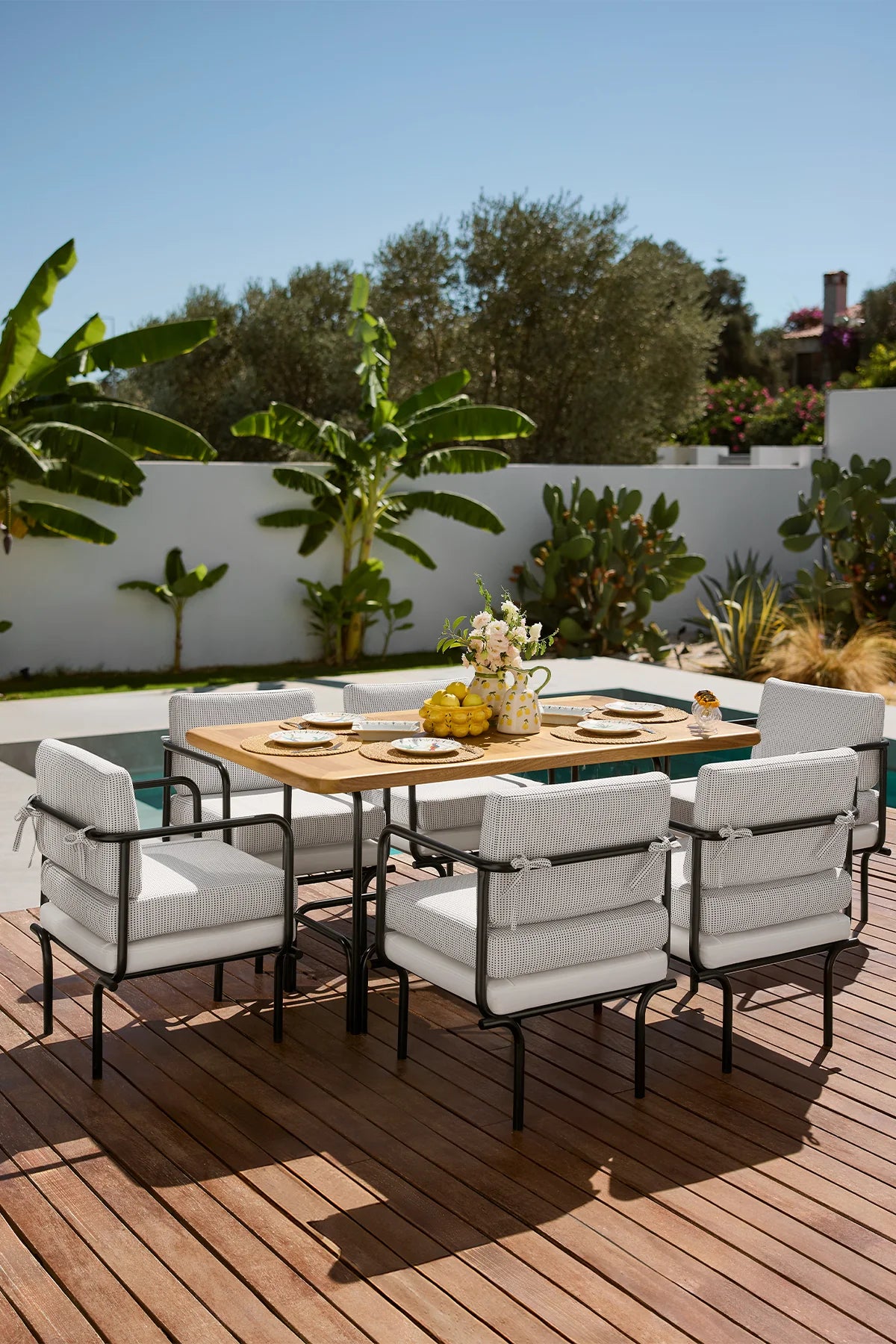 Key West Outdoor Dining Set with black metal chairs, padded outdoor cushions, and a natural iroko wood dining table arranged by the poolside.