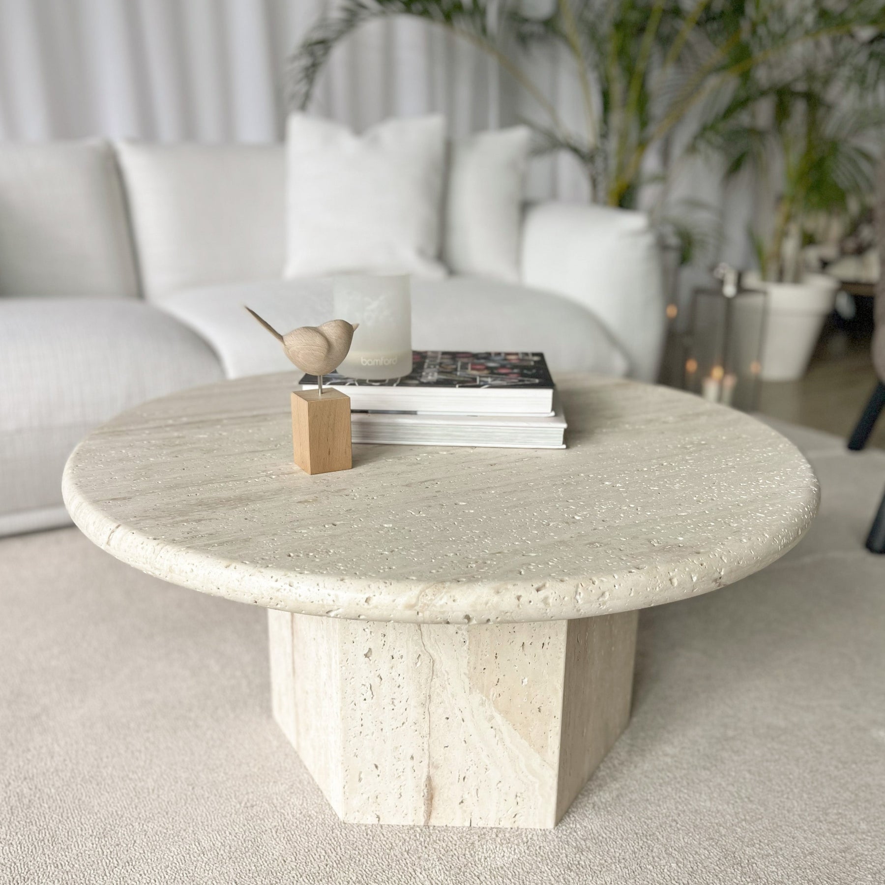 Modern living room featuring a round Luzione travertine coffee table styled with books, candle, and wooden bird figurine; white sofa and plants