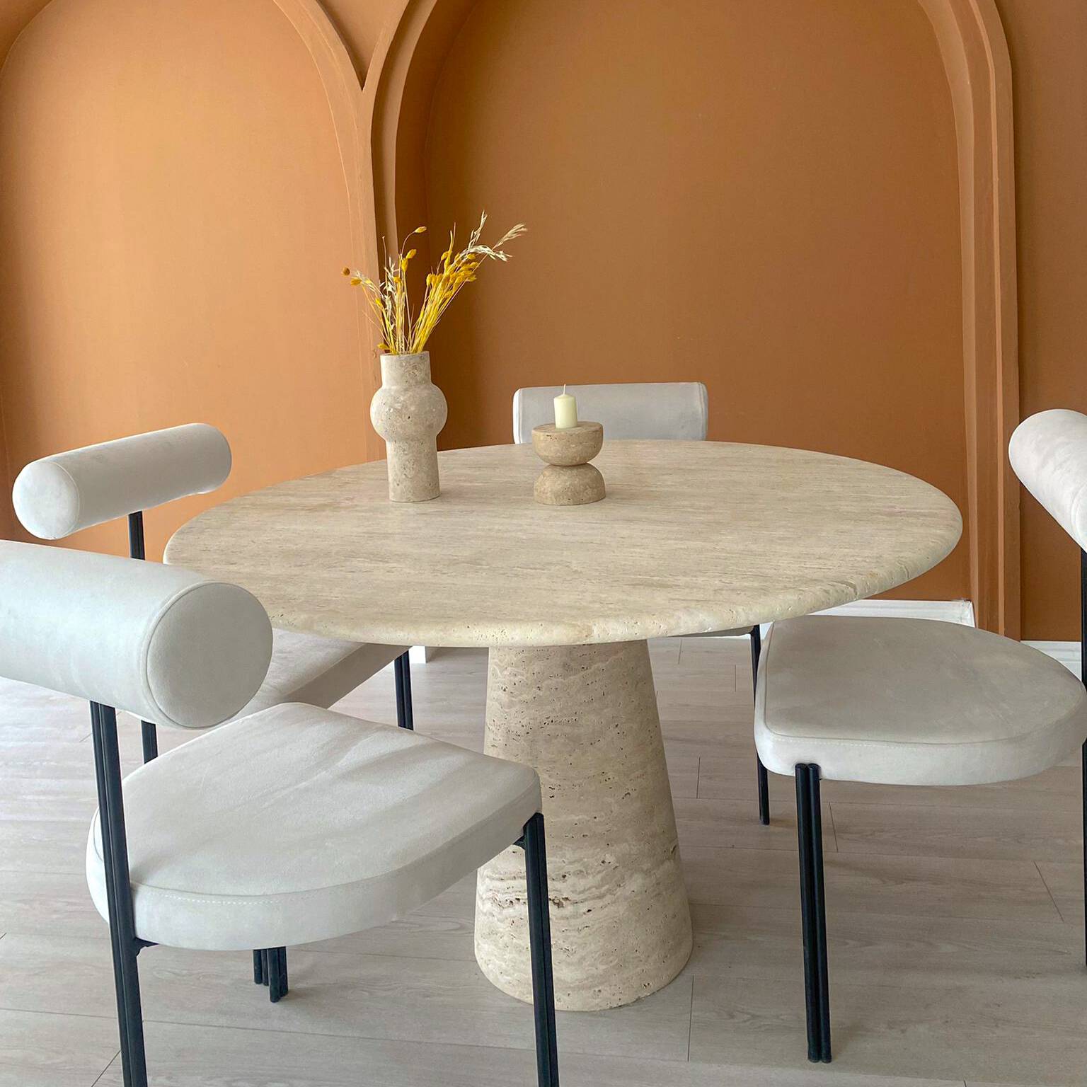 Luzione Round Travertine Dining Table with modern upholstered chairs, dried flowers, candle holder, and earthy-toned wall.