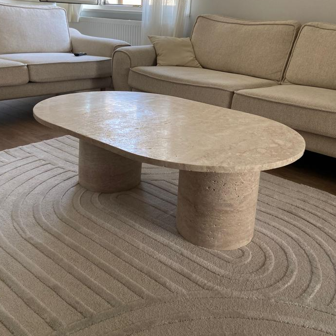 Interior shot of a living room featuring the Luzione Travertine Oval Coffee Table, beige sofas, and a geometric rug.