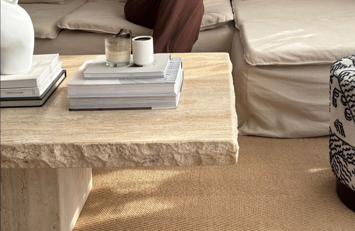 Interior of neutral living room featuring Luzione travertine coffee table styled with books, candles, and a white vase, sofa, ottoman, and area rug