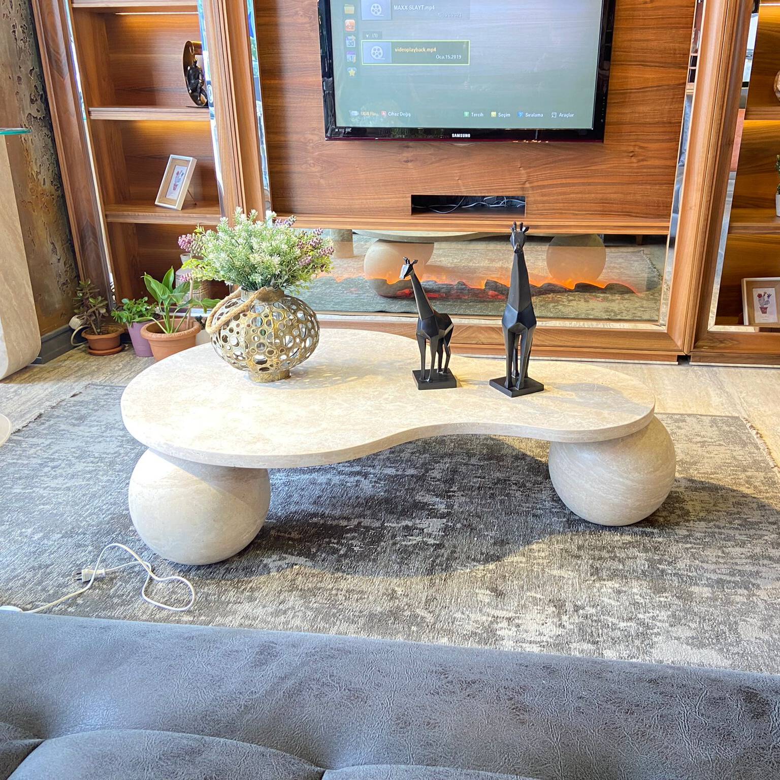 Luzione kidney-shaped travertine coffee table with sphere legs in a modern living room setting with wooden entertainment center