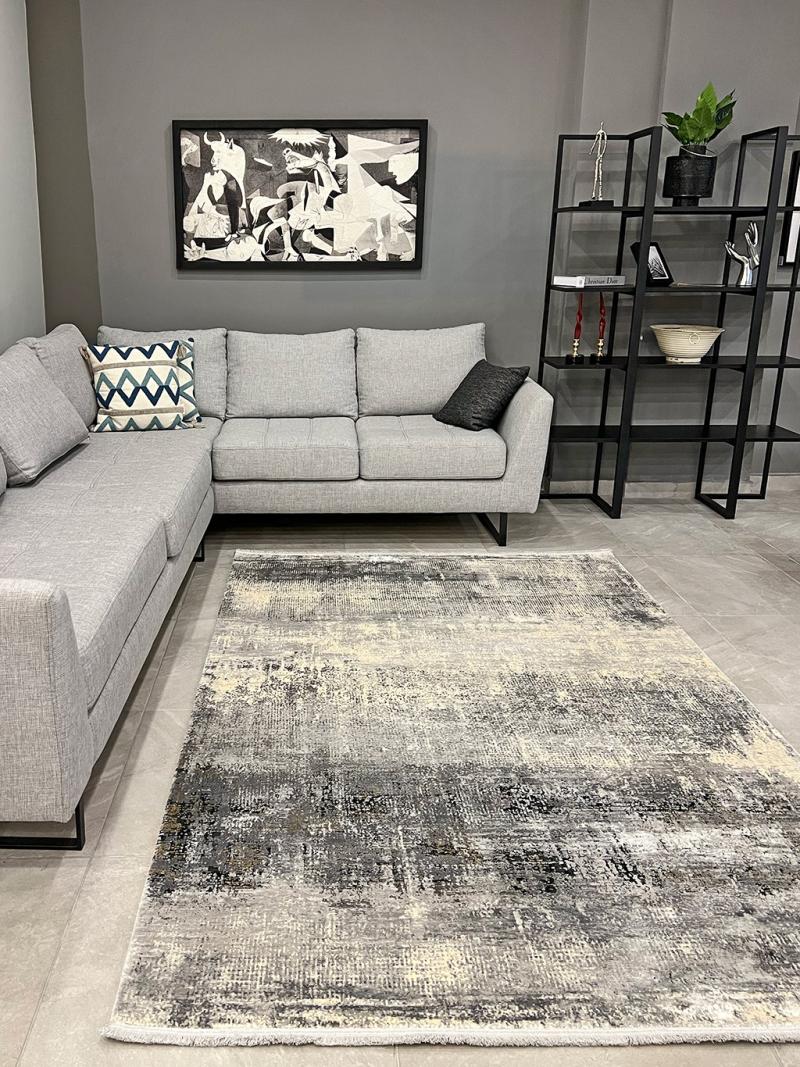 View of modern living room with gray sectional sofa, abstract rug, black shelving. Gray fabric sectional sofa with black legs and decorative pillows. Abstract gray and cream area rug on light gray tile floor. Black metal open shelving unit with various decor items. Black framed abstract wall art above the sofa. Modern interior design, contemporary furniture, living room decor, gray sofa, abstract area rug, open shelving.