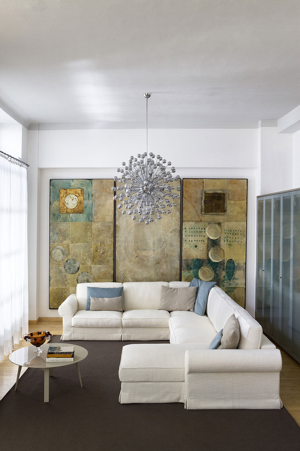 View of living room with white sectional sofa, round table, chandelier, artwork, brown rug, glass cabinet.