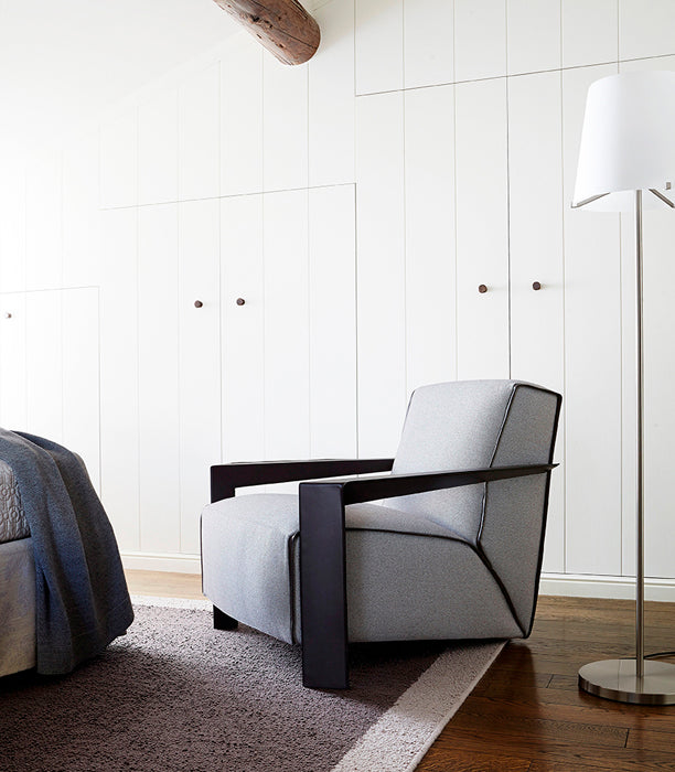 Bedroom view with gray upholstered armchair, floor lamp, and white paneled walls.