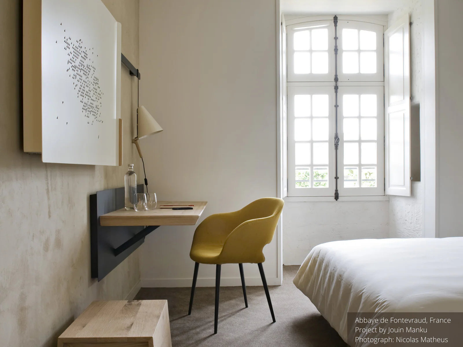 Bedroom with yellow upholstered chair, floating wooden desk, wall art, window.