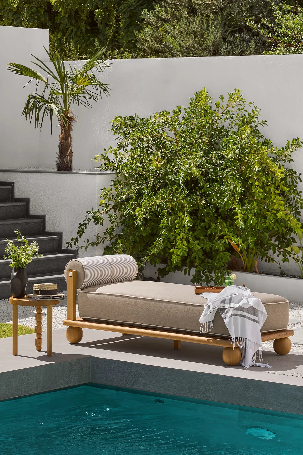 Outdoor lounge chair by a pool with greenery and a white wall in the background