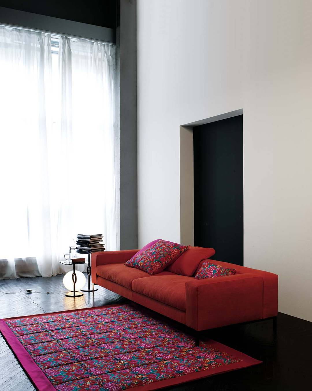 High ceiling living room with red sofa, patterned rug, glass side table, floor lamp, sheer curtains, dark wood floor, white walls, modern furniture, interior design, home decor, contemporary style, luxury seating, colorful textiles, statement rug, minimalist architecture