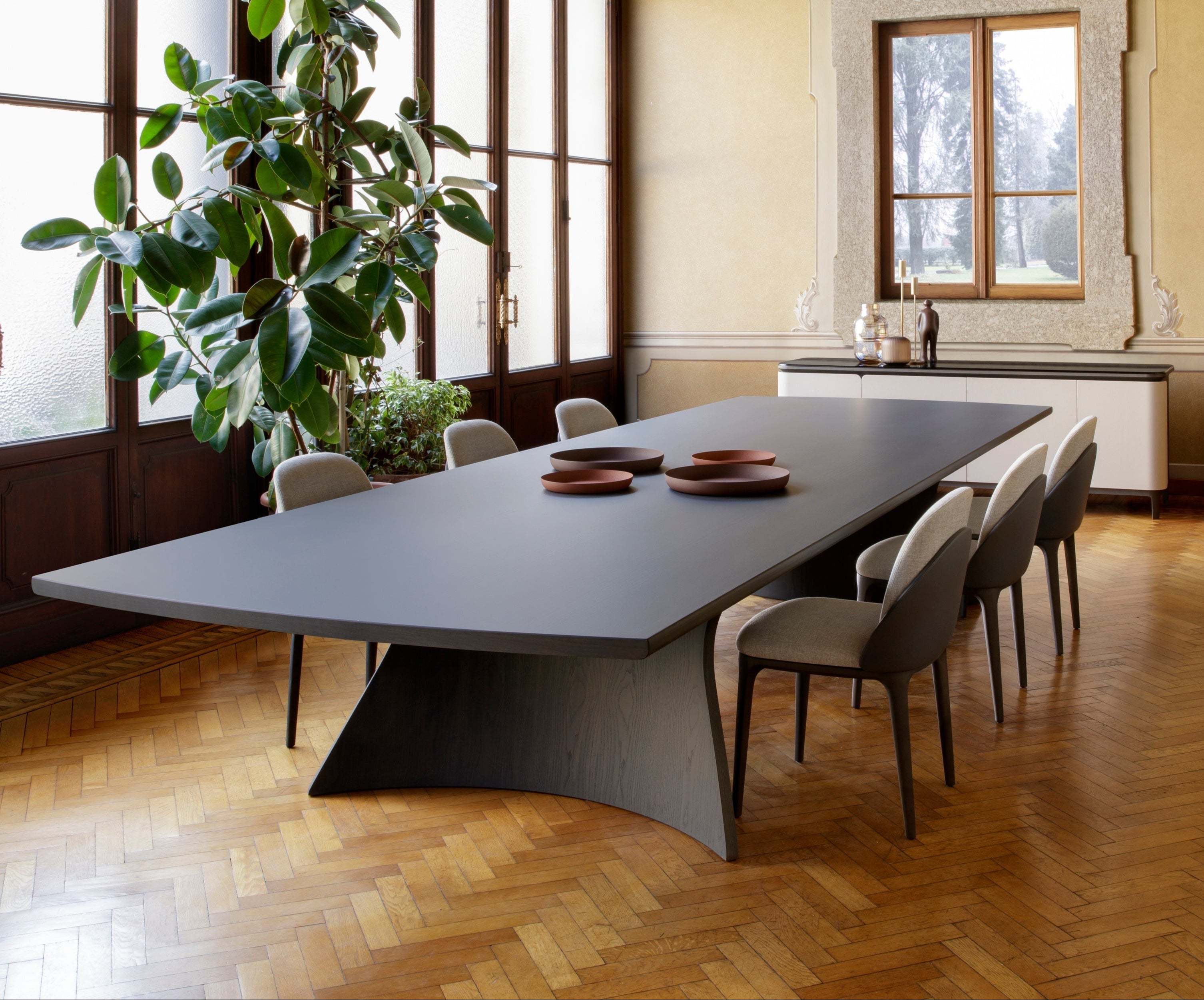 Dining room interior view, long dark dining table, modern chairs, wood floor, tall plant, grandfather clock, sideboard.