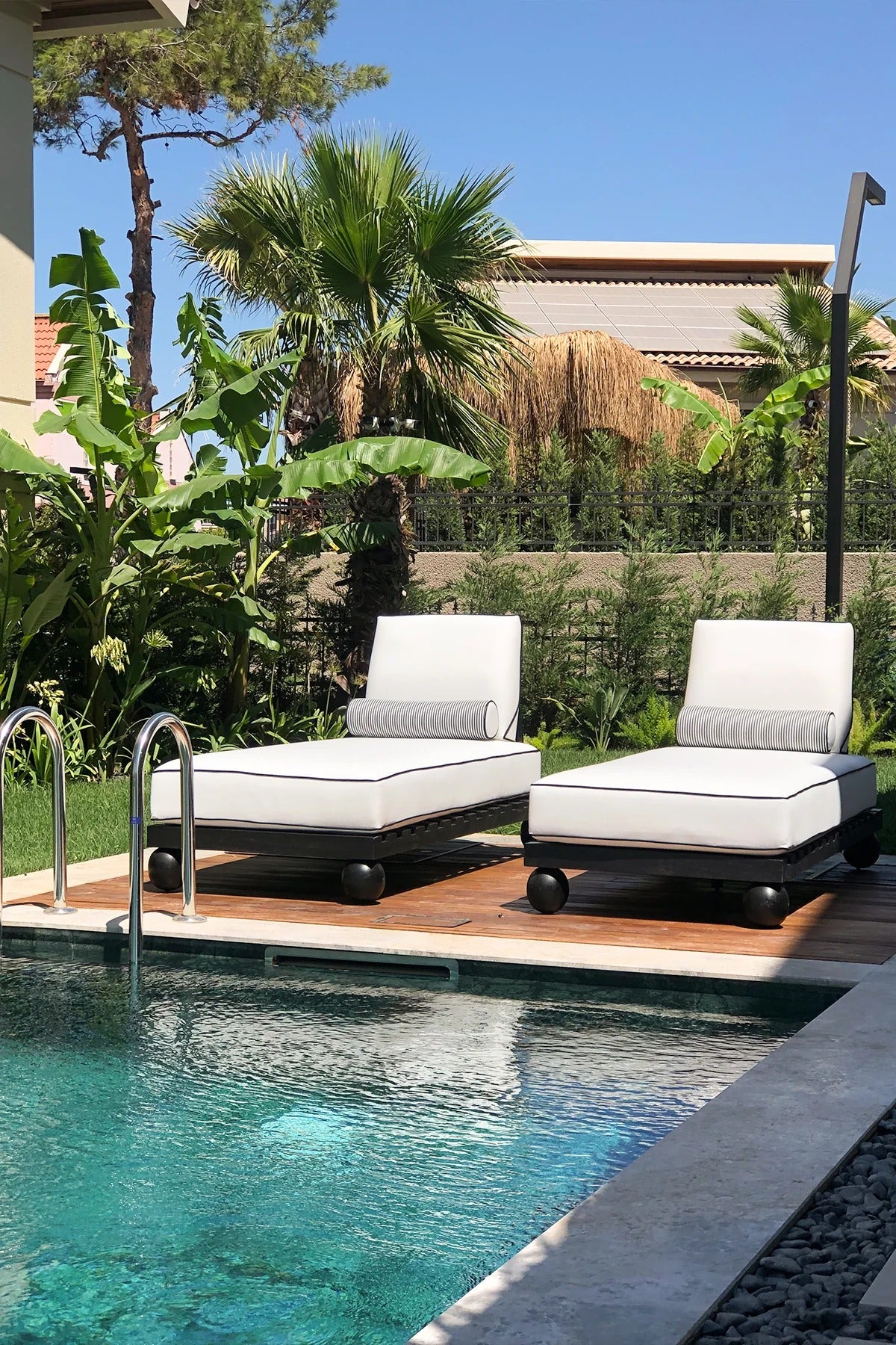 Two white lounge chairs with black bases by a poolside with greenery in the background.
