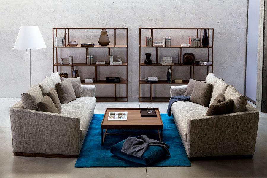 View of two light gray sofas facing each other, dark wood coffee table, blue rug, two dark wood shelving units, and a white floor lamp.