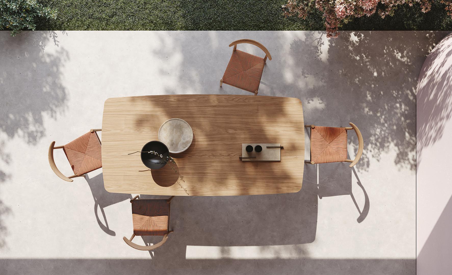  High-angle view of the Casamilano Virgola IN & OUTDOOR dining table with woven chairs on a patio, dappled sunlight, and garden backdrop