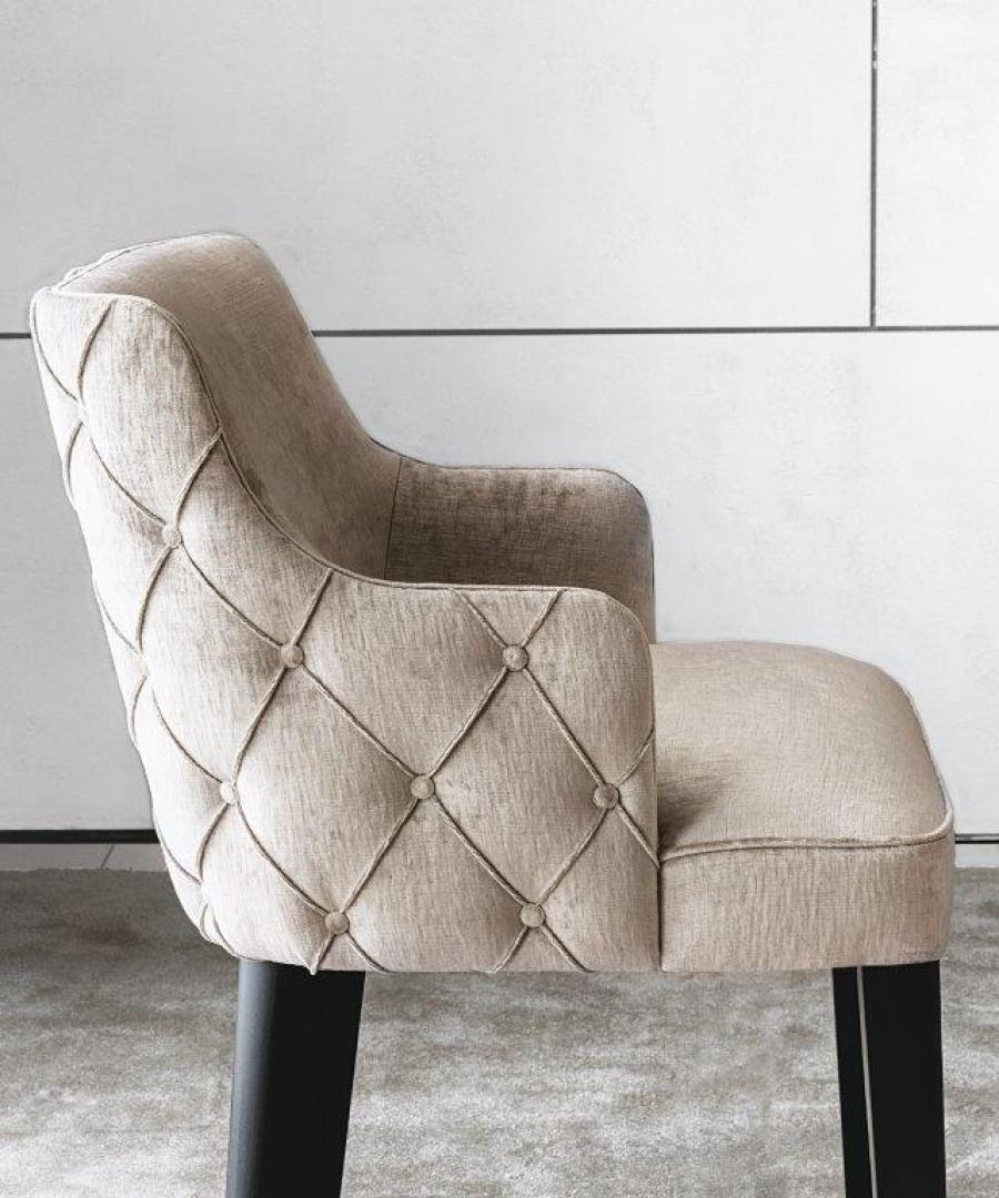  Close-up of the Casamilano Royale Capitonné armchair. Beige upholstery, tufted back, and black legs against a white wall.