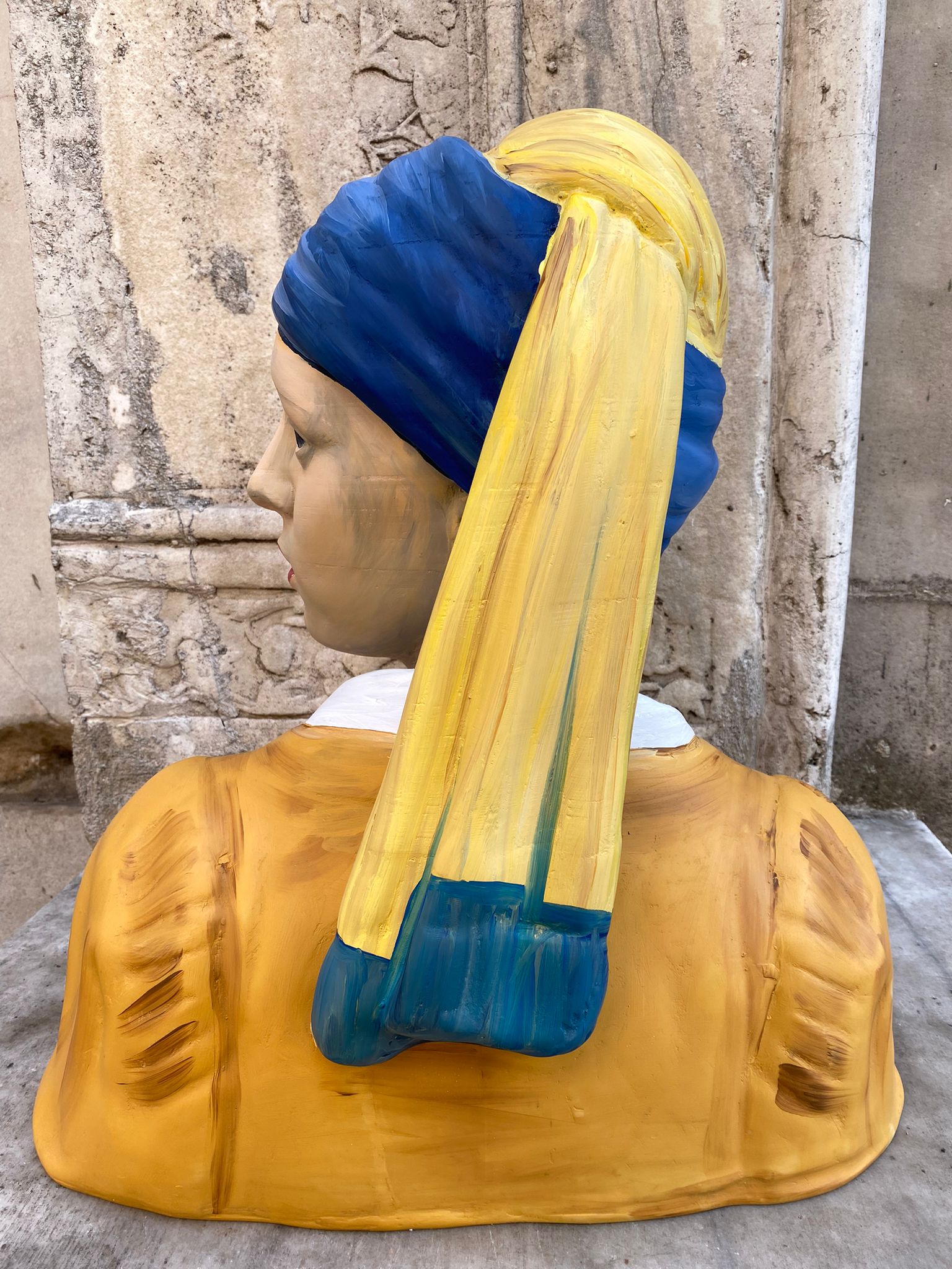 Outdoor view of Girl with a Pearl Earring sculpture, yellow and blue colors, stone wall background, decorative bust, outdoor decor, painted statue, garden sculpture, art reproduction, classic art decor, furniture accent