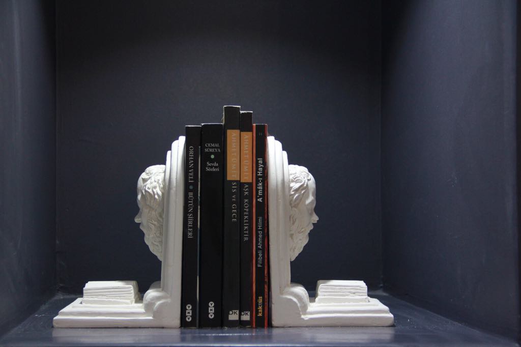 View of white classical bust bookends holding books on a dark shelf. 