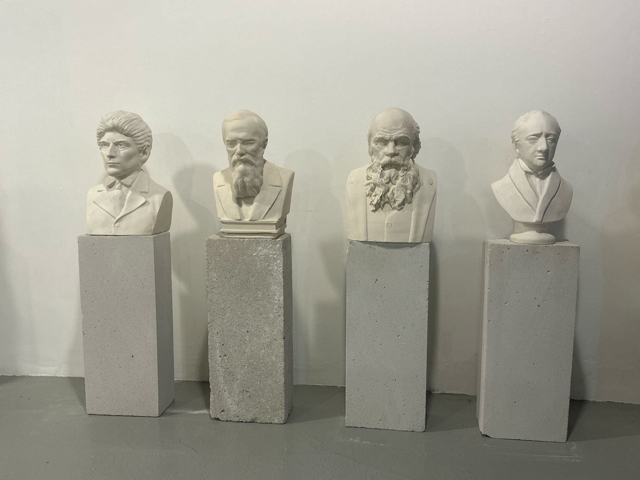 Four white busts on concrete pedestals against white wall.