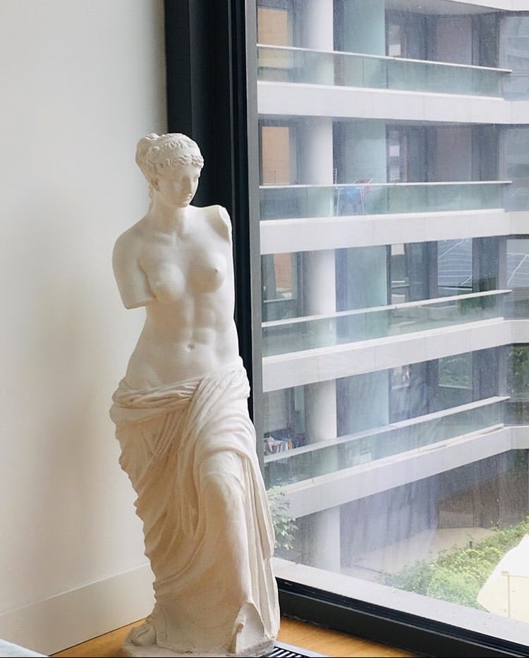 White female torso sculpture near window overlooking modern building balconies.