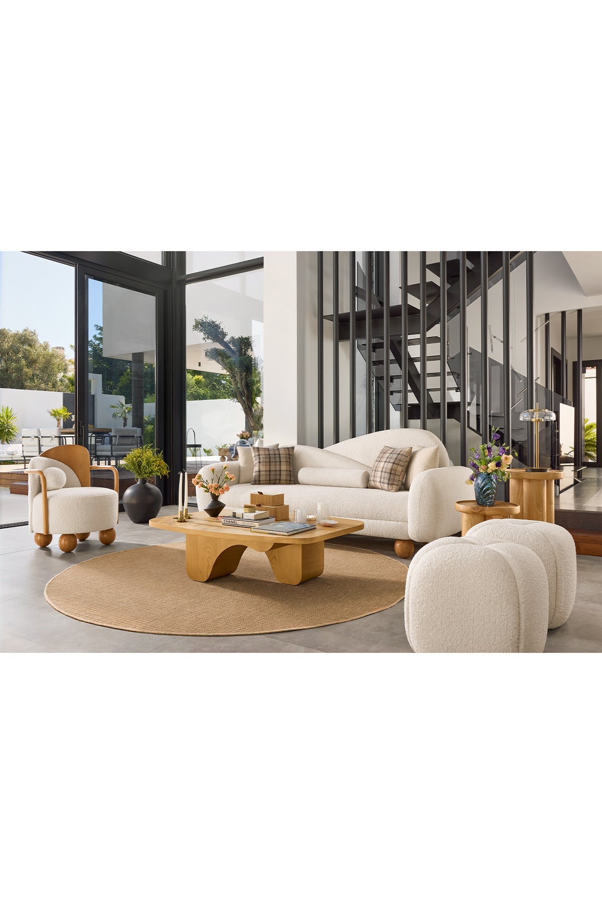 Modern living room with white boucle sofa, armchair, wooden coffee table, round rug, ottomans, glass doors, and staircase.
