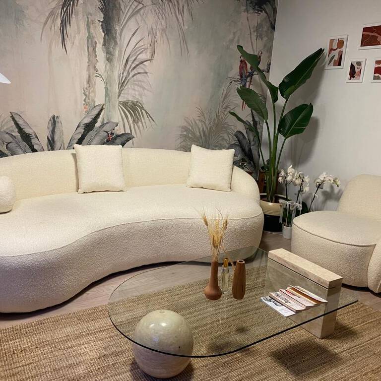 Stylish living room featuring a Luzione cream bouclé curved sofa, armchair, glass coffee table, jute rug, and tropical wallpaper for a modern, inviting space