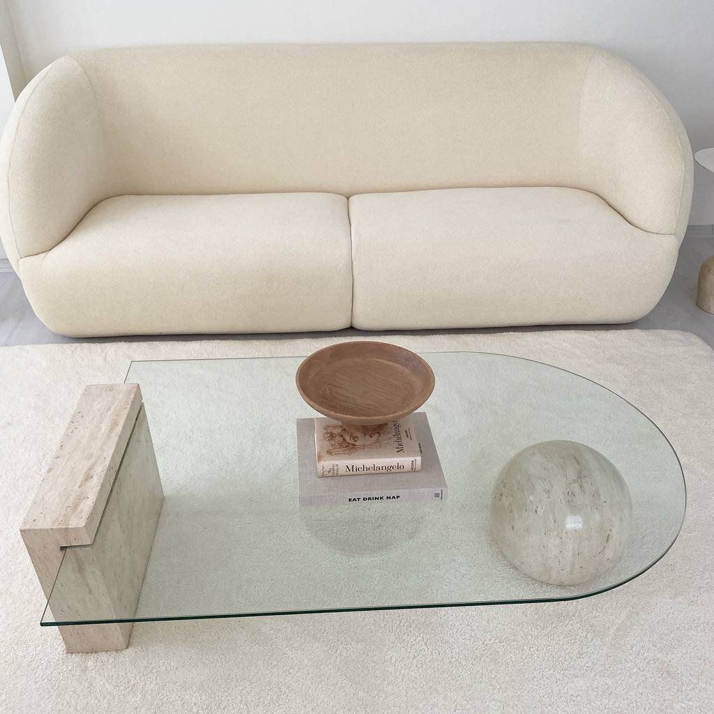 Modern living room featuring a cream curved sofa, glass-topped Luzione coffee table with travertine base, and plush white rug