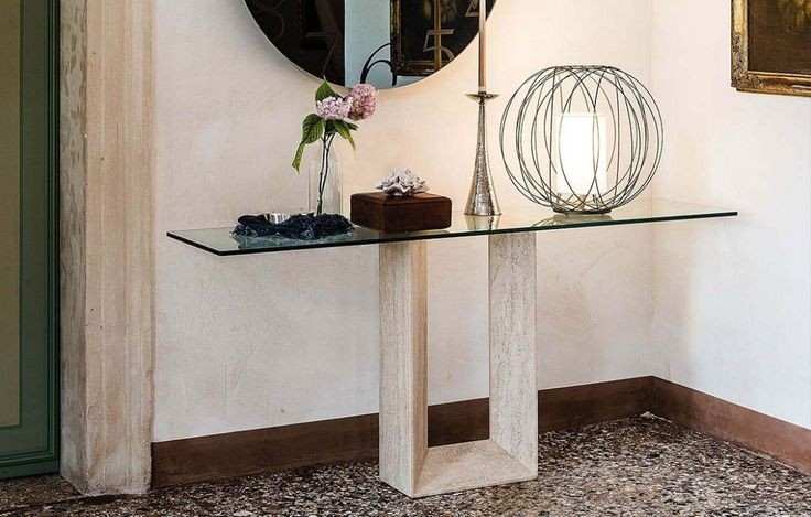 A modern console table with a clear glass top and U-shaped travertine base, adorned with elegant decor, perfect for a contemporary entryway