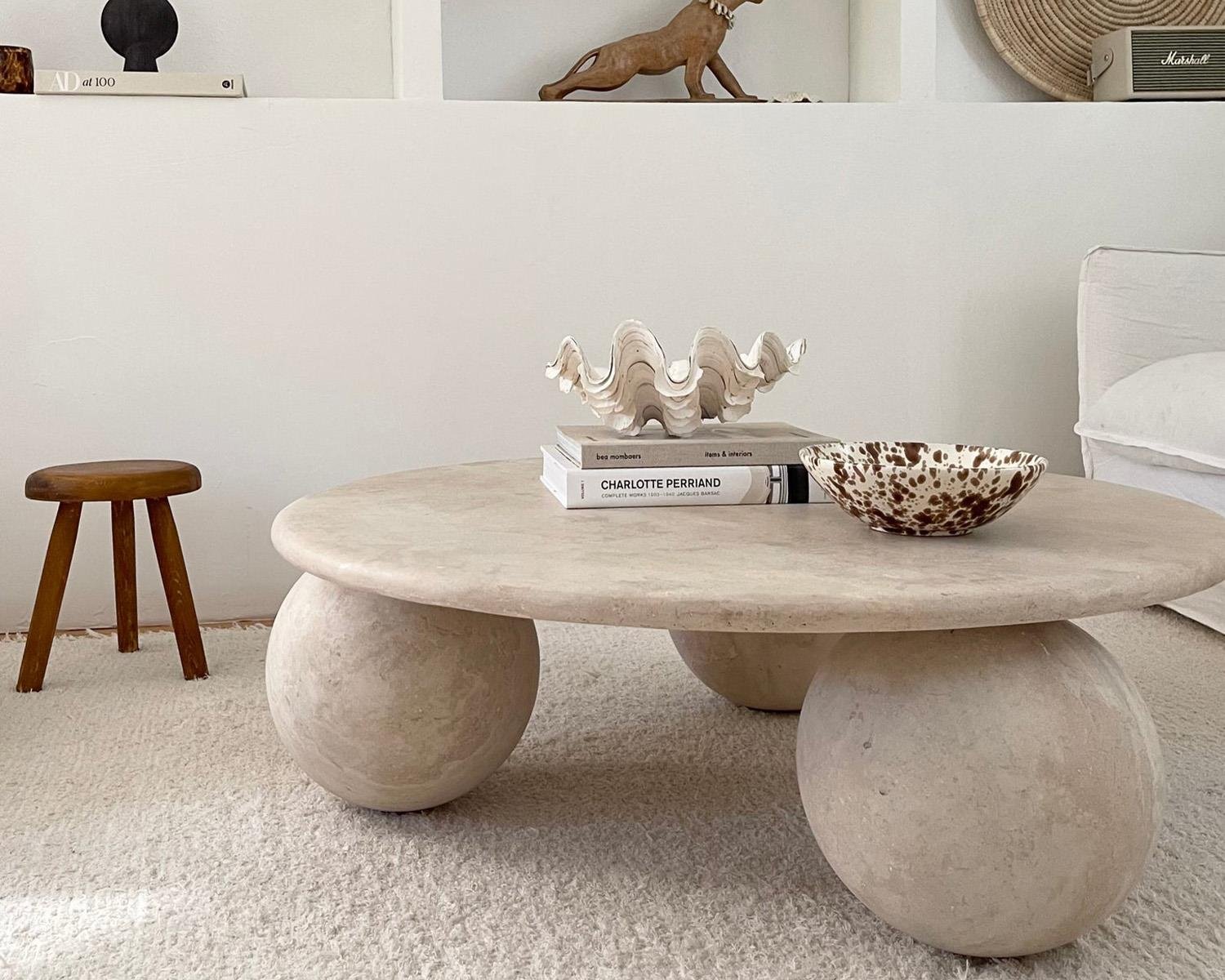 View of living room with round travertine coffee table and wood stool. Travertine coffee table with three spherical legs. White clam shell decor, spotted bowl, and books on table. White shaggy rug on floor.
