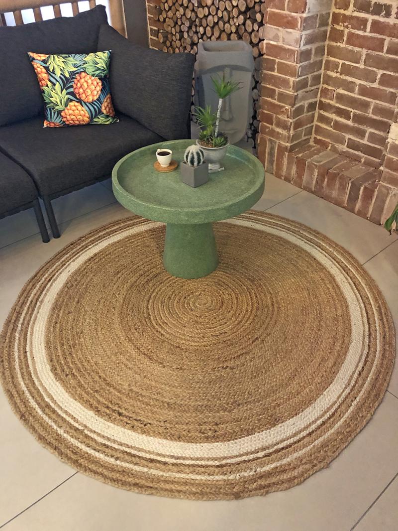 Interior view of a living room featuring a round jute rug, green side table, modern sofa, and indoor plants, creating a cozy atmosphere