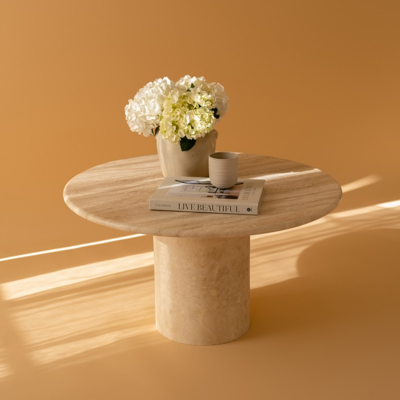 Eye-level shot of Luzione Cylinder Leg Travertine Coffee Table with hydrangeas, mug, and book on a tan background. Modern living room furniture