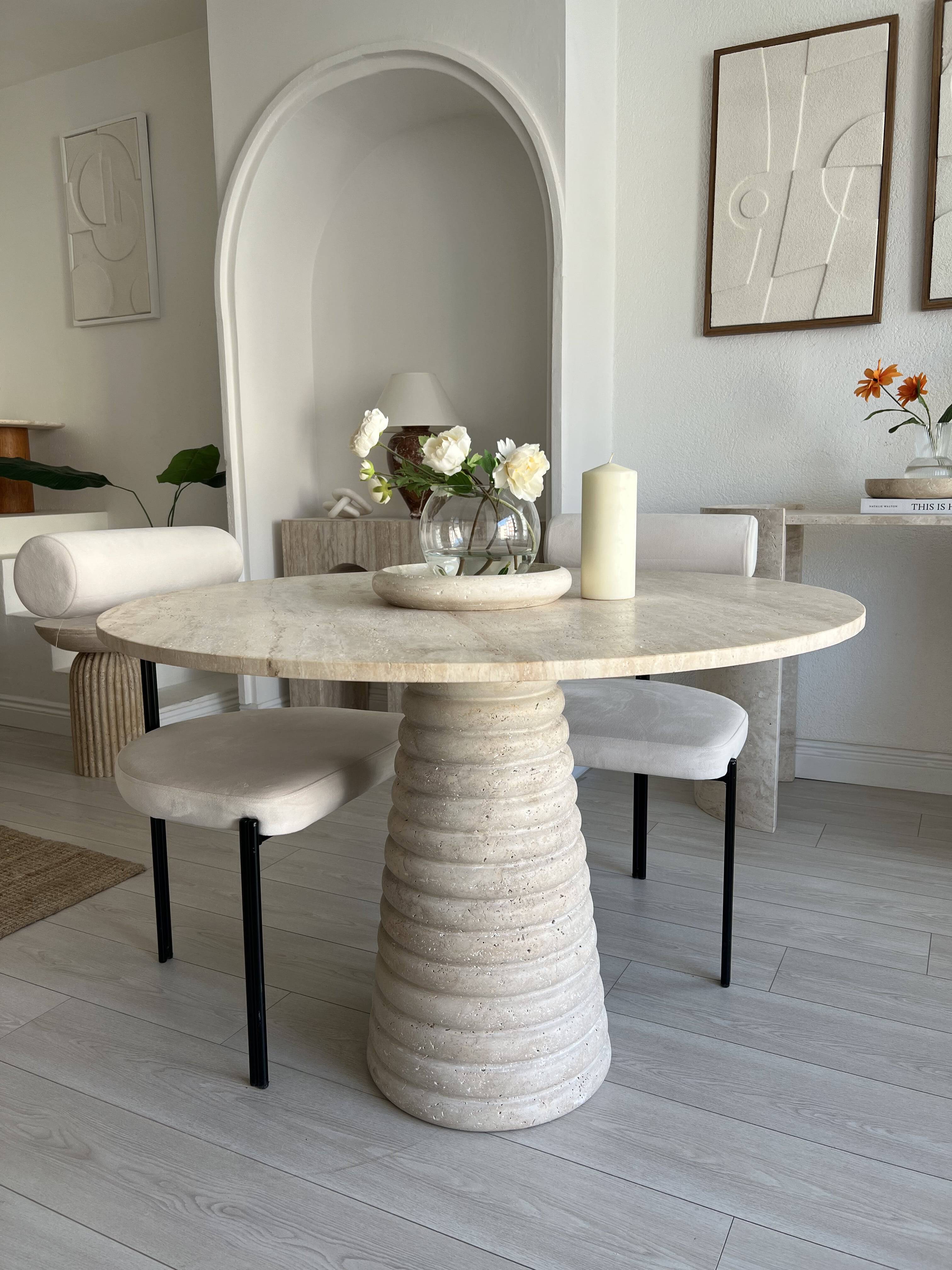 View of dining area with round travertine table and chairs, light flooring, arched niche, and wall art.
travertine dining table with round top and ribbed pedestal base.
Dining table with round travertine top and ribbed base, two upholstered chairs.
White upholstered dining chairs with black metal legs next to travertine table.
White wall with arched niche and two framed abstract relief wall art pieces.
White flowers in glass vase and large candle on round travertine table.
Light gray wood plank flooring und