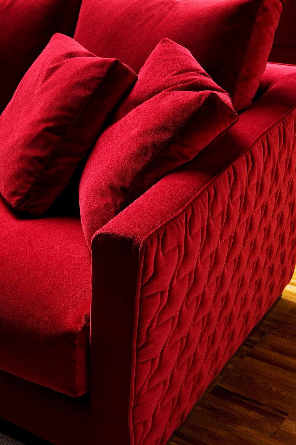 Close up view of red velvet sofa with quilted side panel and cushions on wood floor, modern furniture, living room seating.