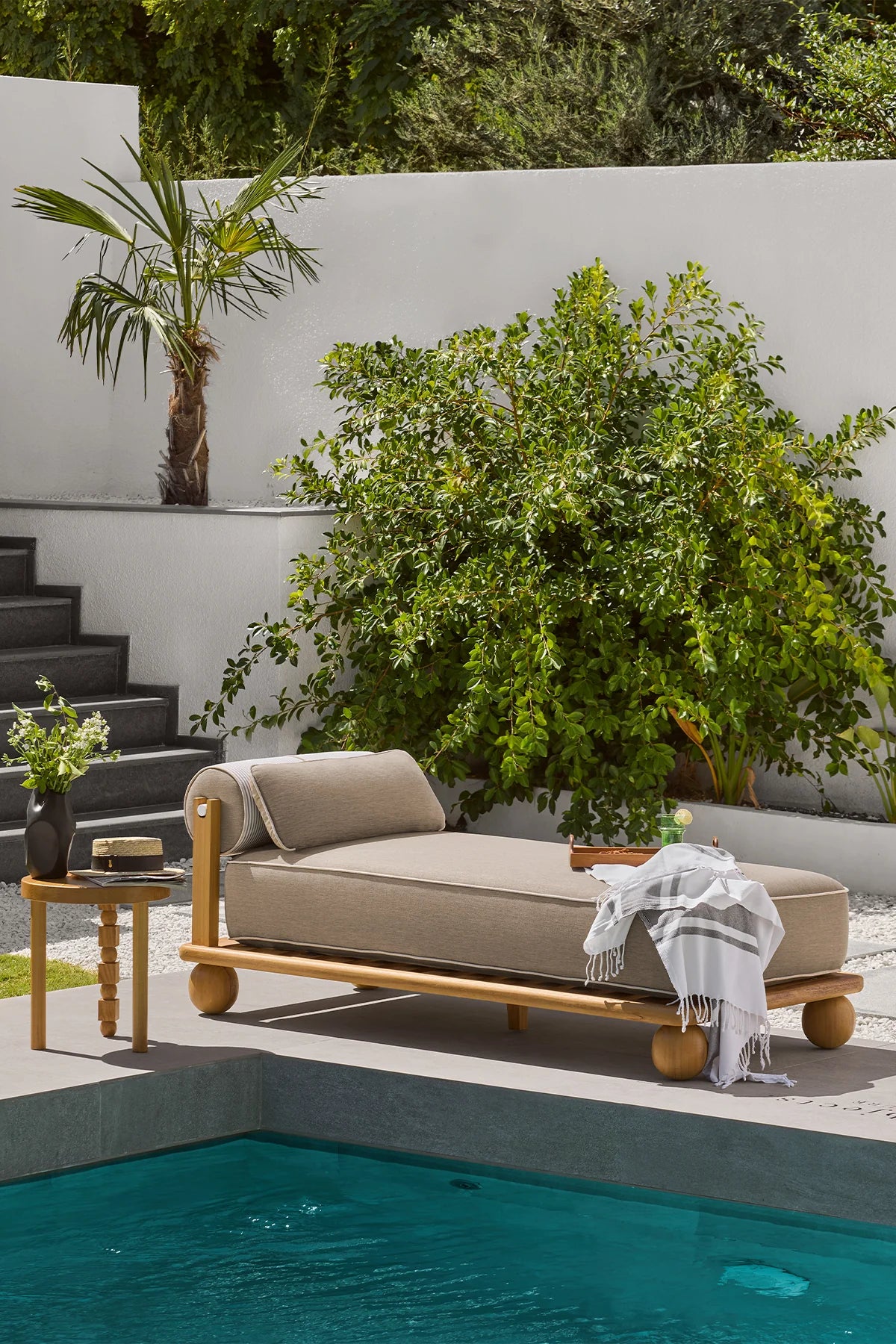Natural-finish iroko wood sun lounger with beige outdoor cushion placed beside a pool.