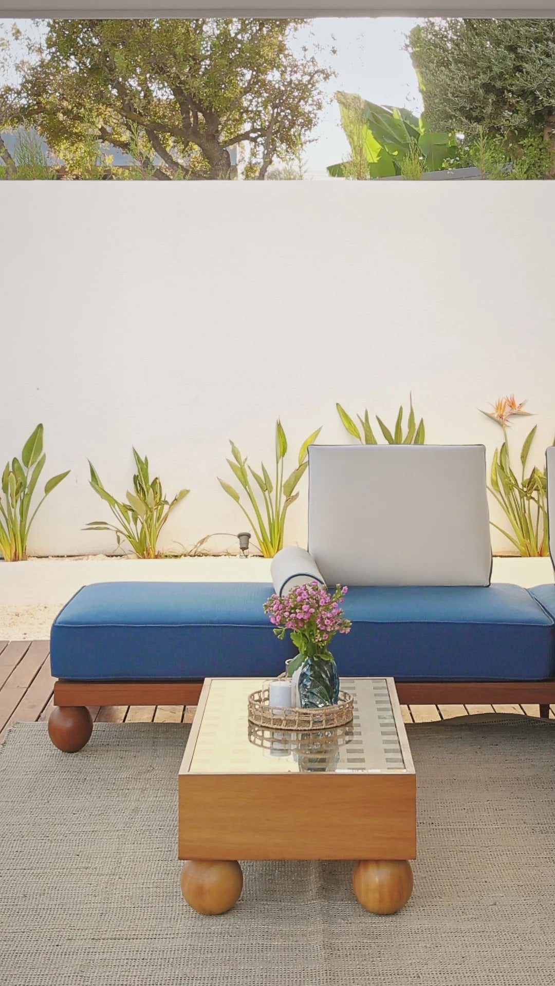 Outdoor seating area with a blue cushioned bench and wooden coffee table featuring a vase of flowers.