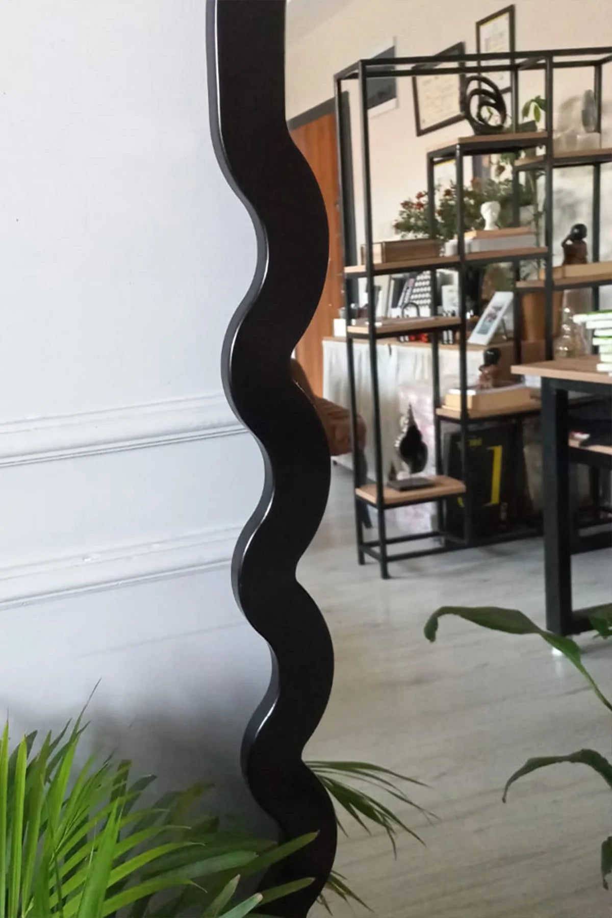 Black wavy mirror against a white wall with indoor plants and furniture in the background